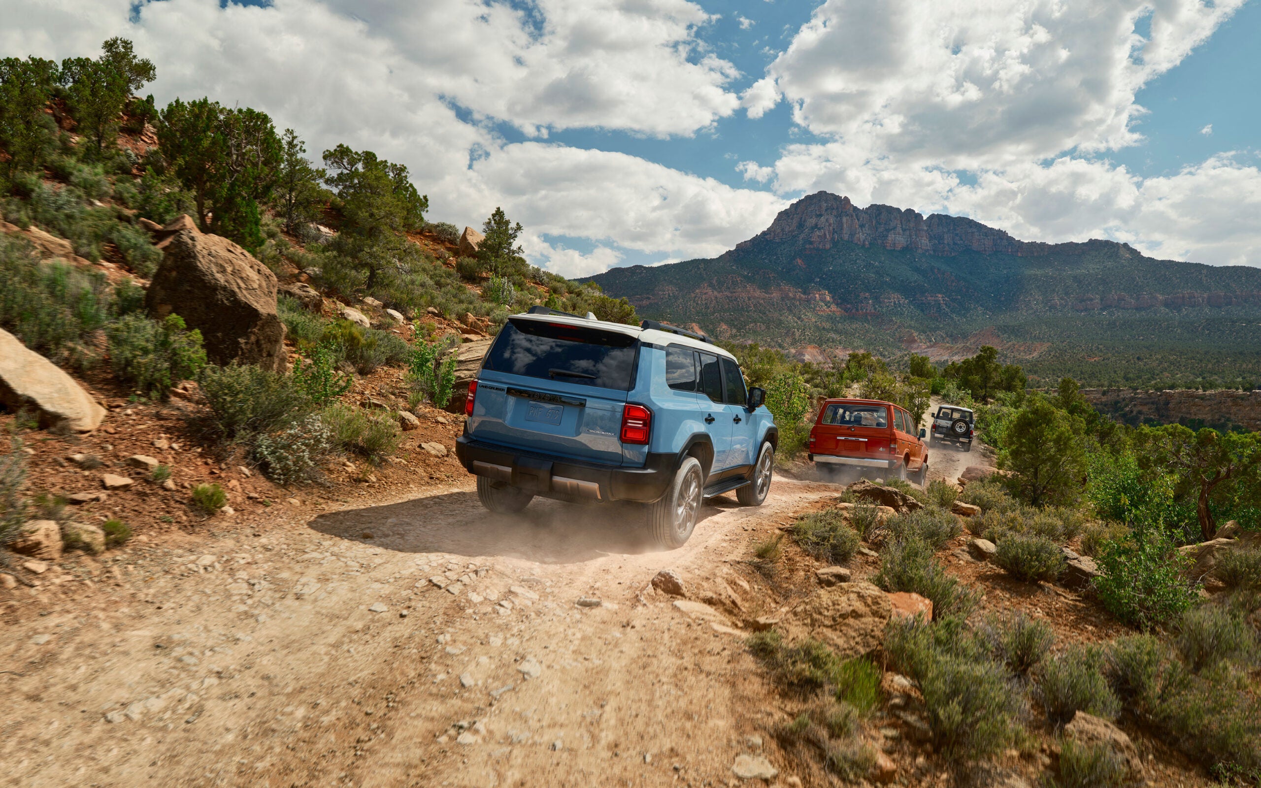 A blue 2024 Toyota Land Cruiser J250 follows two classic Land Cruisers down an unpaved desert road.